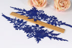 a close-up view of a blue lace fabric with intricate floral patterns, placed on a white surface. A wooden ruler is positioned in the center of the fabric, likely used for measuring or cutting the lace. In the background, there are two pink roses, adding a touch of color to the scene.