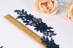 a close-up view of a blue lace piece with intricate floral designs, placed next to a wooden ruler on a white surface. In the background, there are two pink roses, adding a touch of color to the scene.