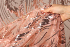A hand is holding a piece of fabric with a pink and black sequin pattern, against a background of a floral pattern.