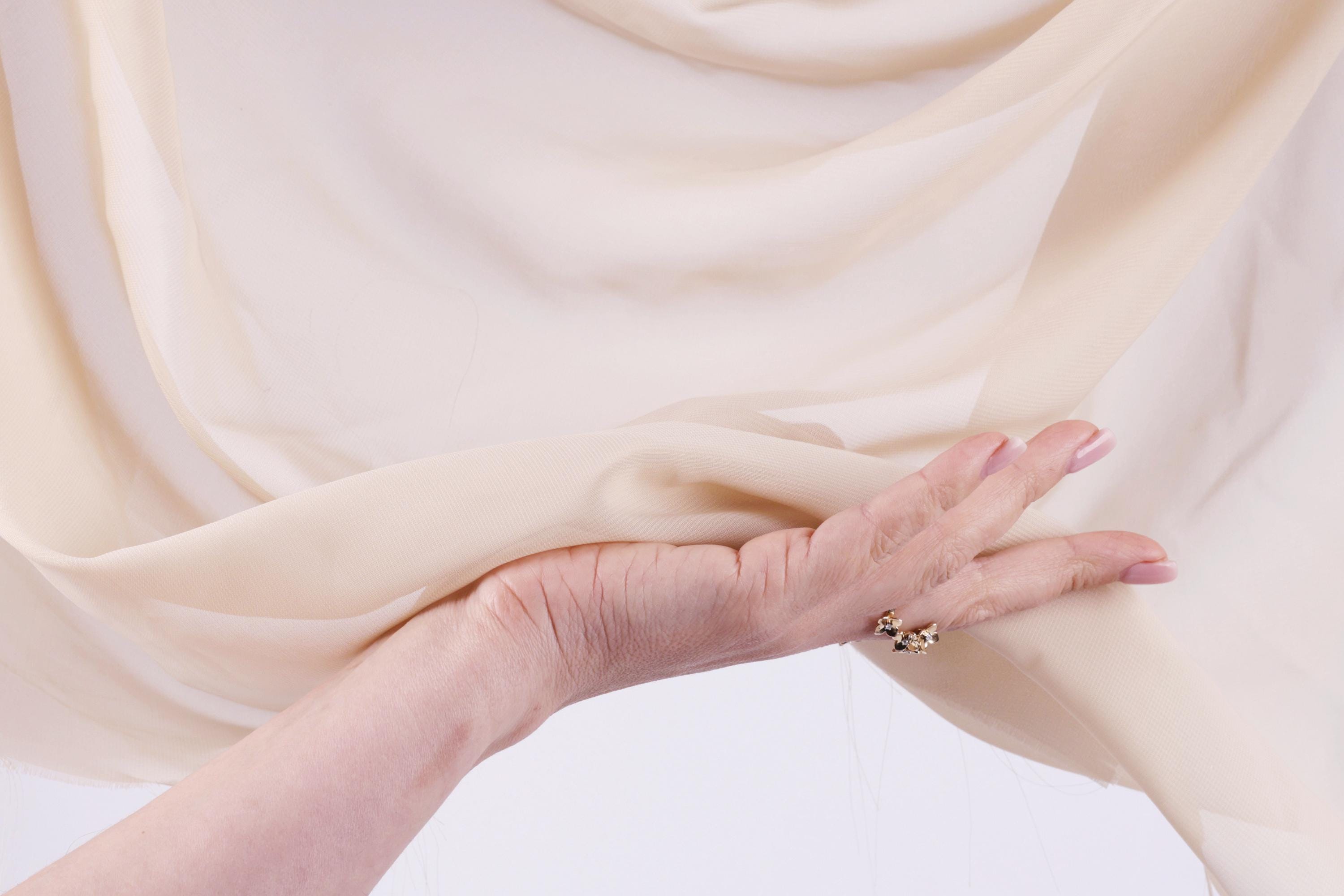 A hand is holding a piece of light-colored fabric, possibly a sheer or sheer-like material, with a ring visible on one of the fingers.
