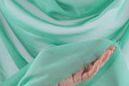 A hand is holding a piece of green fabric, which appears to be a sheer or sheer-like material. The fabric has a soft, flowing texture and is draped over the hand, creating a sense of movement and elegance.
