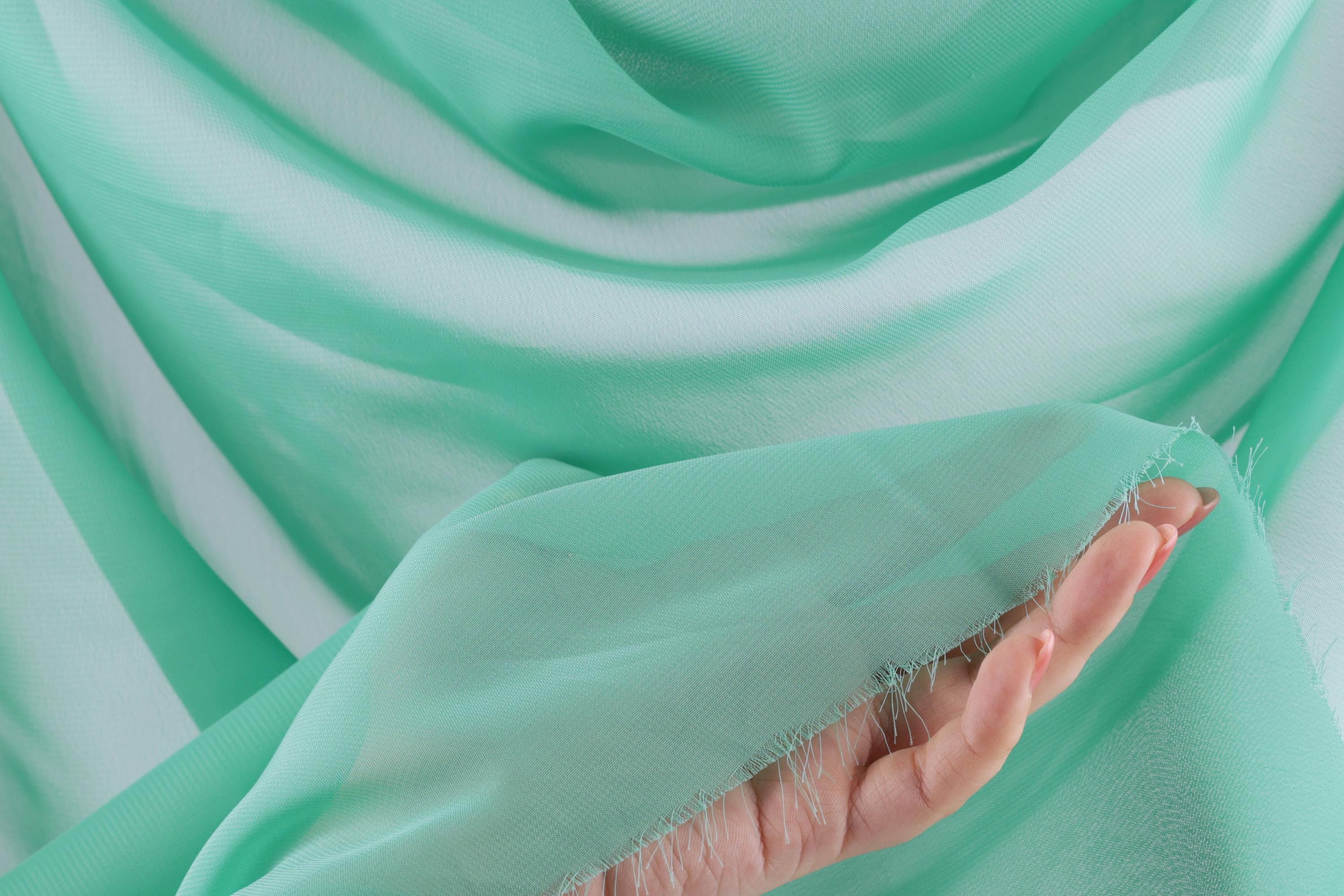 A hand is holding a piece of green fabric, which appears to be a sheer or sheer-like material. The fabric has a soft, flowing texture and is draped over the hand, creating a sense of movement and elegance.