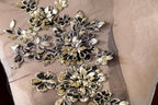 a close-up view of a delicate, intricate floral embroidery on a fabric surface. The embroidery is composed of numerous small, black and gold flowers arranged in a visually appealing pattern. The flowers are intricately detailed, showcasing the skill and craftsmanship of the embroiderer. The fabric appears to be a light-colored material, possibly a sheer or sheer-like fabric, which complements the delicate appearance of the flowers.