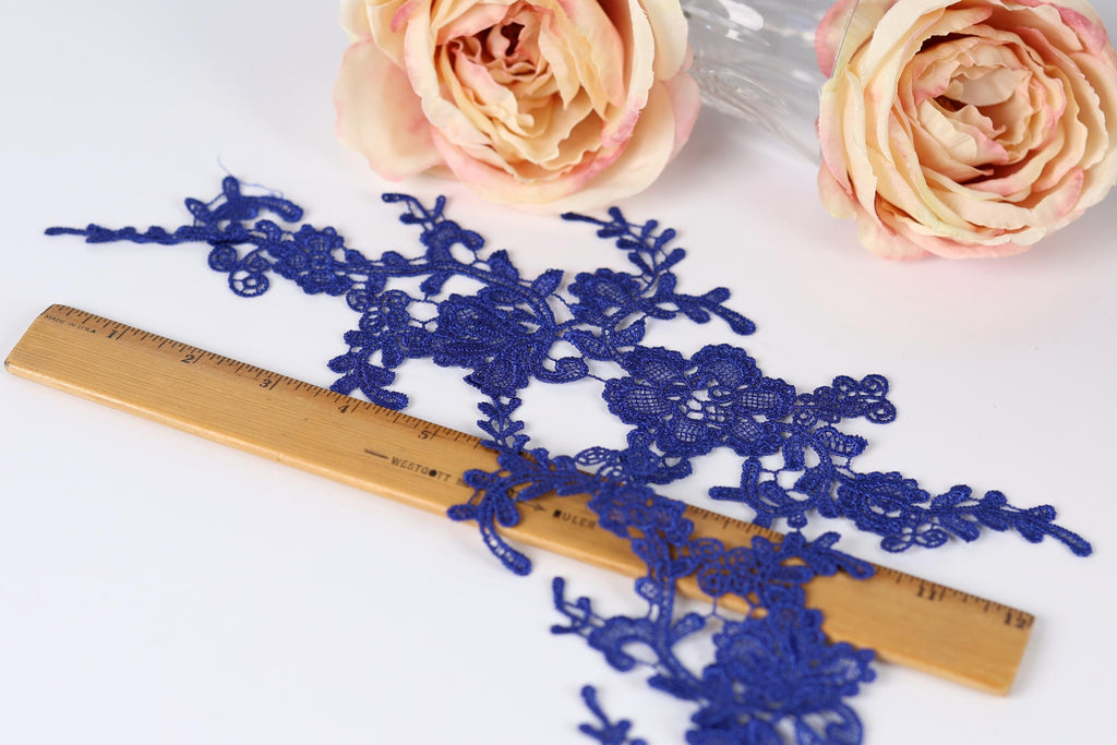 a close-up view of a blue lace fabric with intricate floral patterns, placed on a wooden ruler for size reference. In the background, there are two pink roses, adding a touch of color to the scene.
