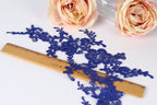 a close-up view of a blue lace fabric with intricate floral patterns, placed on a wooden ruler for size reference. In the background, there are two pink roses, adding a touch of color to the scene.