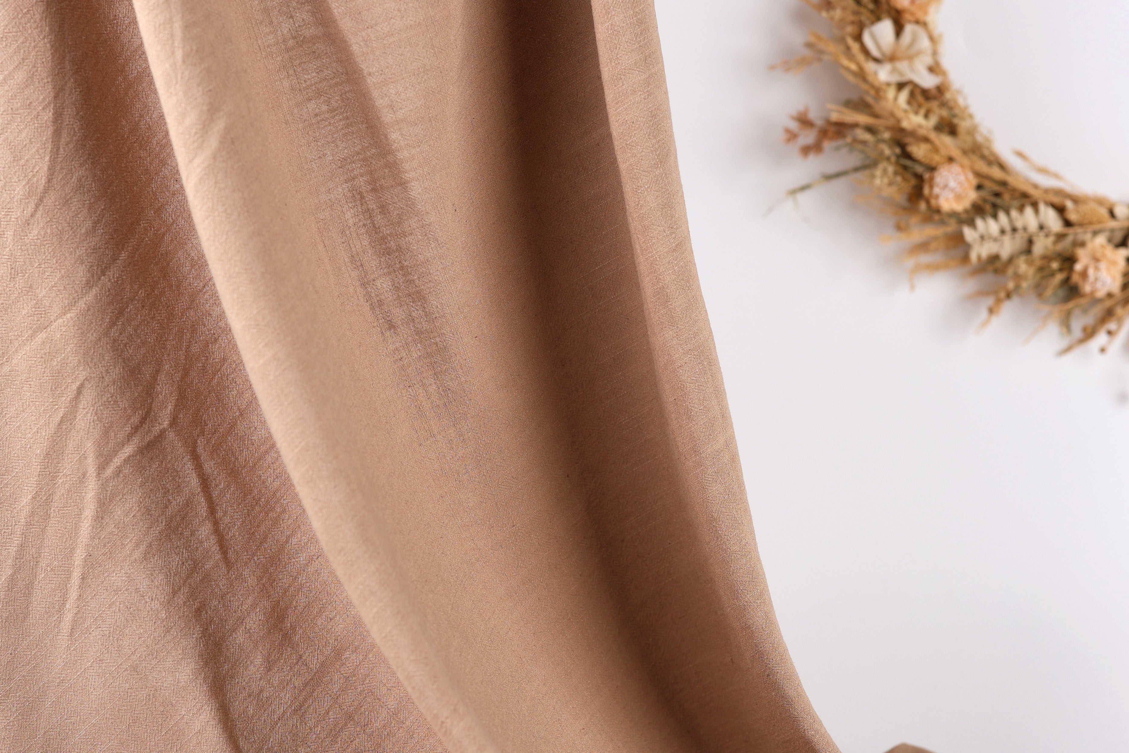 a close-up view of a beige fabric, possibly a curtain or textile, with a wreath of dried flowers or branches placed next to it on a white background.