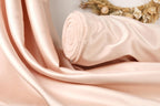 A close-up image of a soft, light pink satin fabric, possibly a fabric or material, with a curled edge and a floral arrangement in the background.