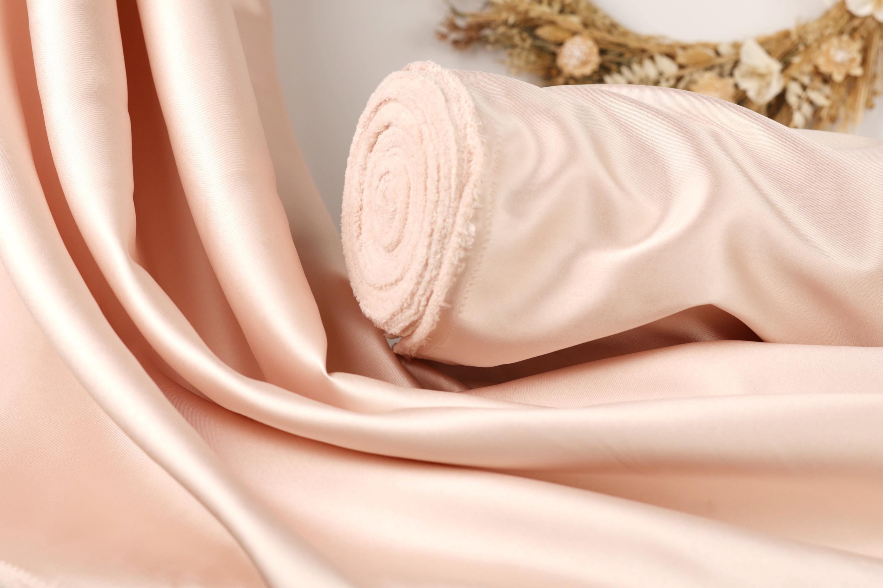 A close-up image of a soft, light pink satin fabric, possibly a fabric or material, with a curled edge and a floral arrangement in the background.