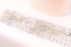 a close-up view of a delicate, intricate piece of jewelry, possibly a brooch or a necklace, with numerous small, sparkling crystals or beads attached to it. The jewelry piece is placed on a transparent ruler, which is lying on a white surface.