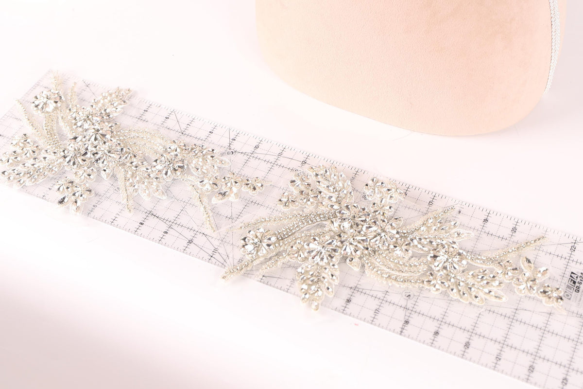 a close-up view of a delicate, intricate piece of jewelry, possibly a brooch or a necklace, with numerous small, sparkling crystals or beads attached to it. The jewelry piece is placed on a transparent ruler, which is lying on a white surface.