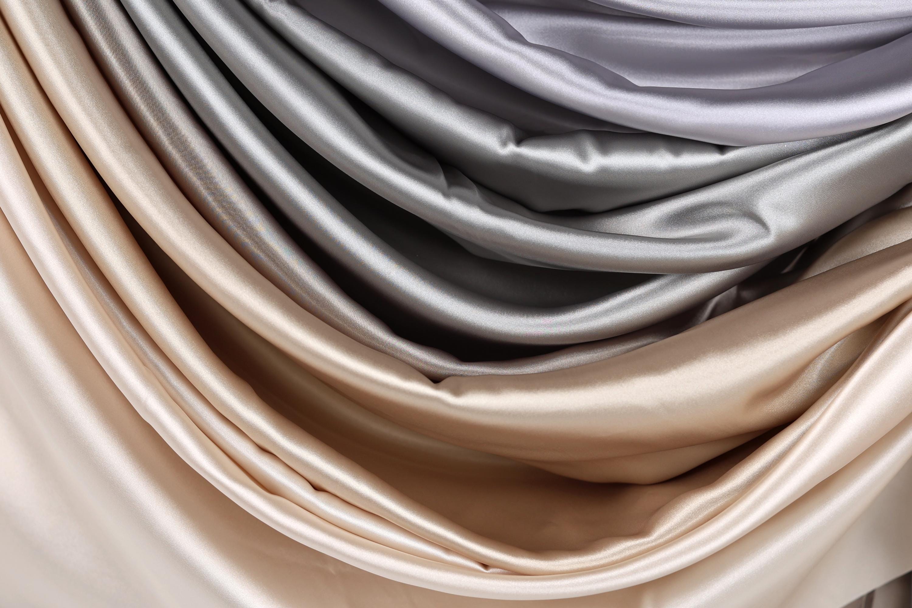 a close-up view of a pile of fabric, with a gradient of colors ranging from light to dark shades of gray and beige. The fabric appears to be made of satin or a similar smooth, shiny material.