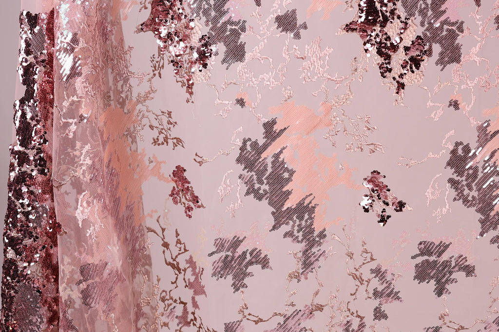 a close-up view of a fabric with a pink and black floral pattern. The fabric appears to be made of sequin material, with a mix of pink and black sequins visible throughout the design. The pattern is intricate and detailed, showcasing the craftsmanship of the fabric.