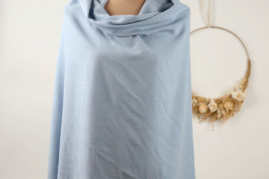 A mannequin wearing a light blue draped garment, with a wreath of dried flowers hanging on the wall behind it.
