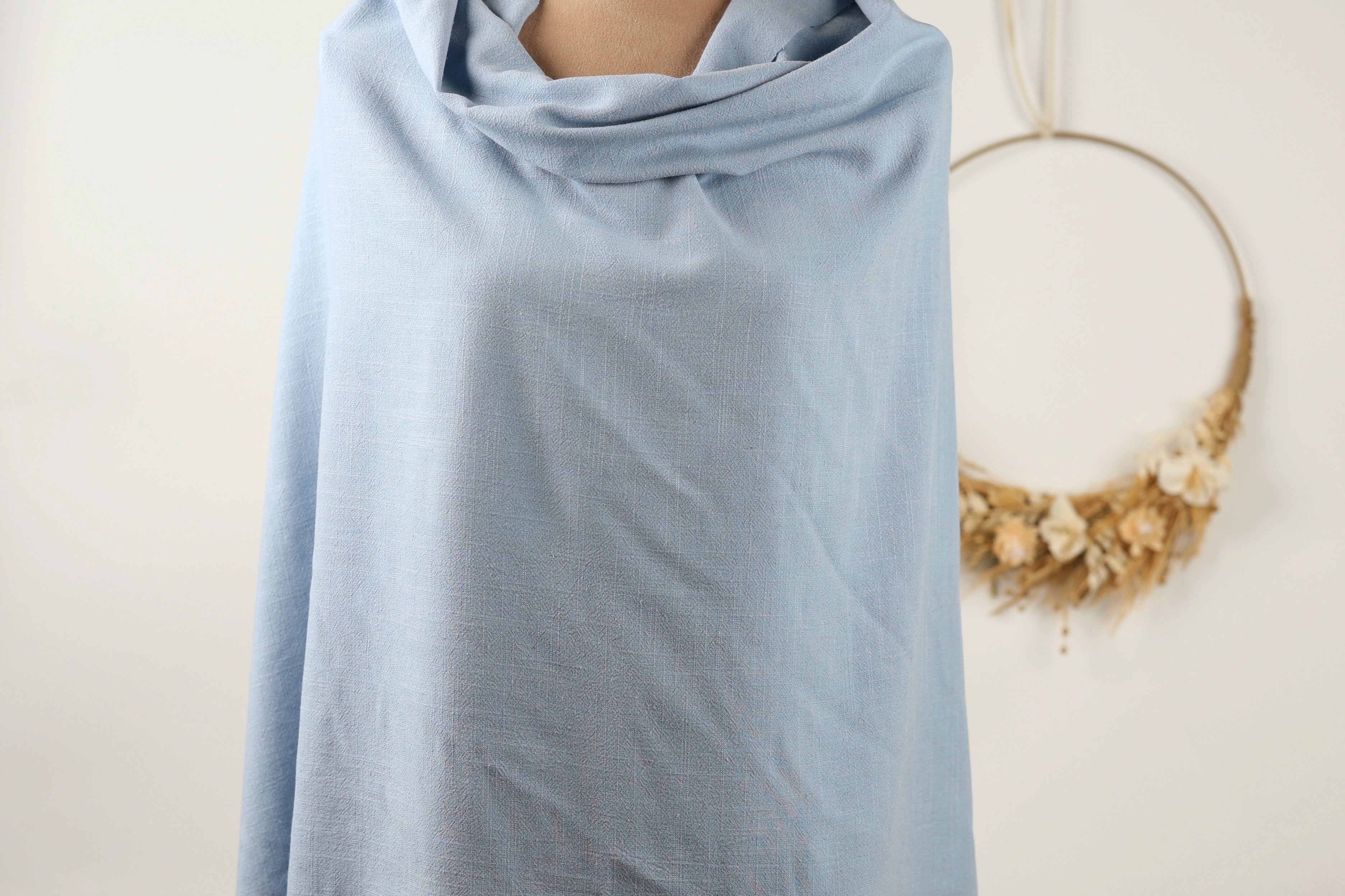 A mannequin wearing a light blue draped garment, with a wreath of dried flowers hanging on the wall behind it.