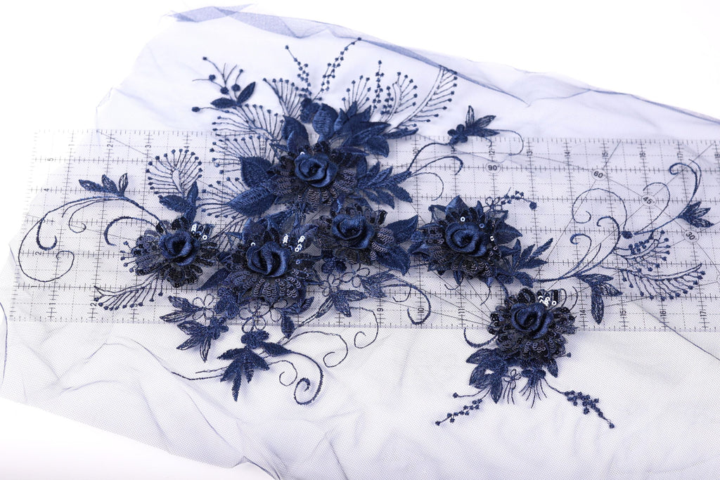a close-up view of a delicate blue floral embroidery on a white background. The embroidery is composed of various blue flowers and leaves, creating a visually appealing and intricate design.