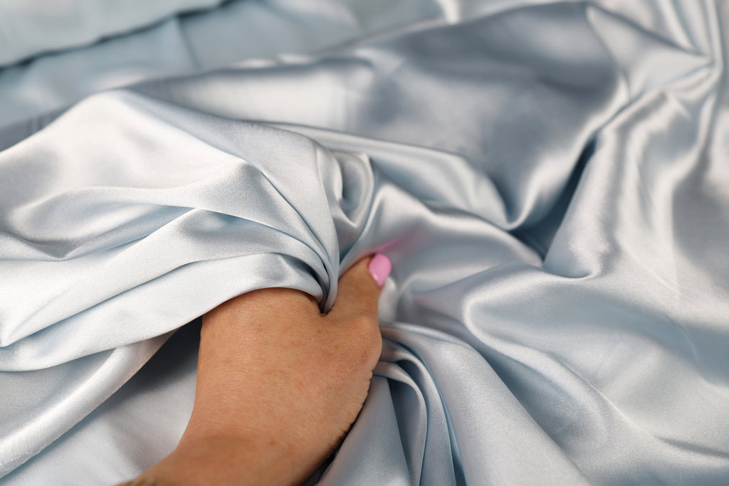 A hand with pink nail polish rests on a crumpled, gray satin sheet.