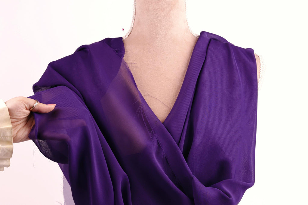 A hand is holding a purple fabric, possibly a dress, against a white background.