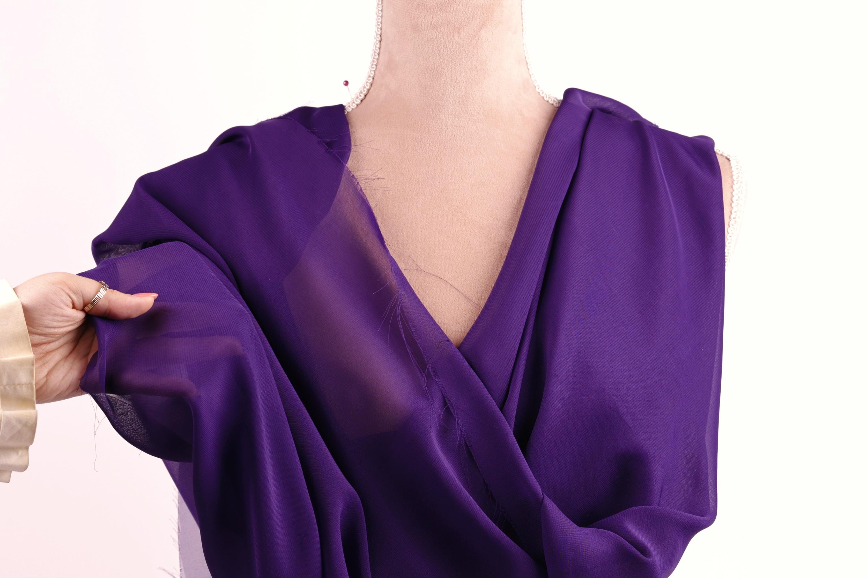 A hand is holding a purple fabric, possibly a dress, against a white background.