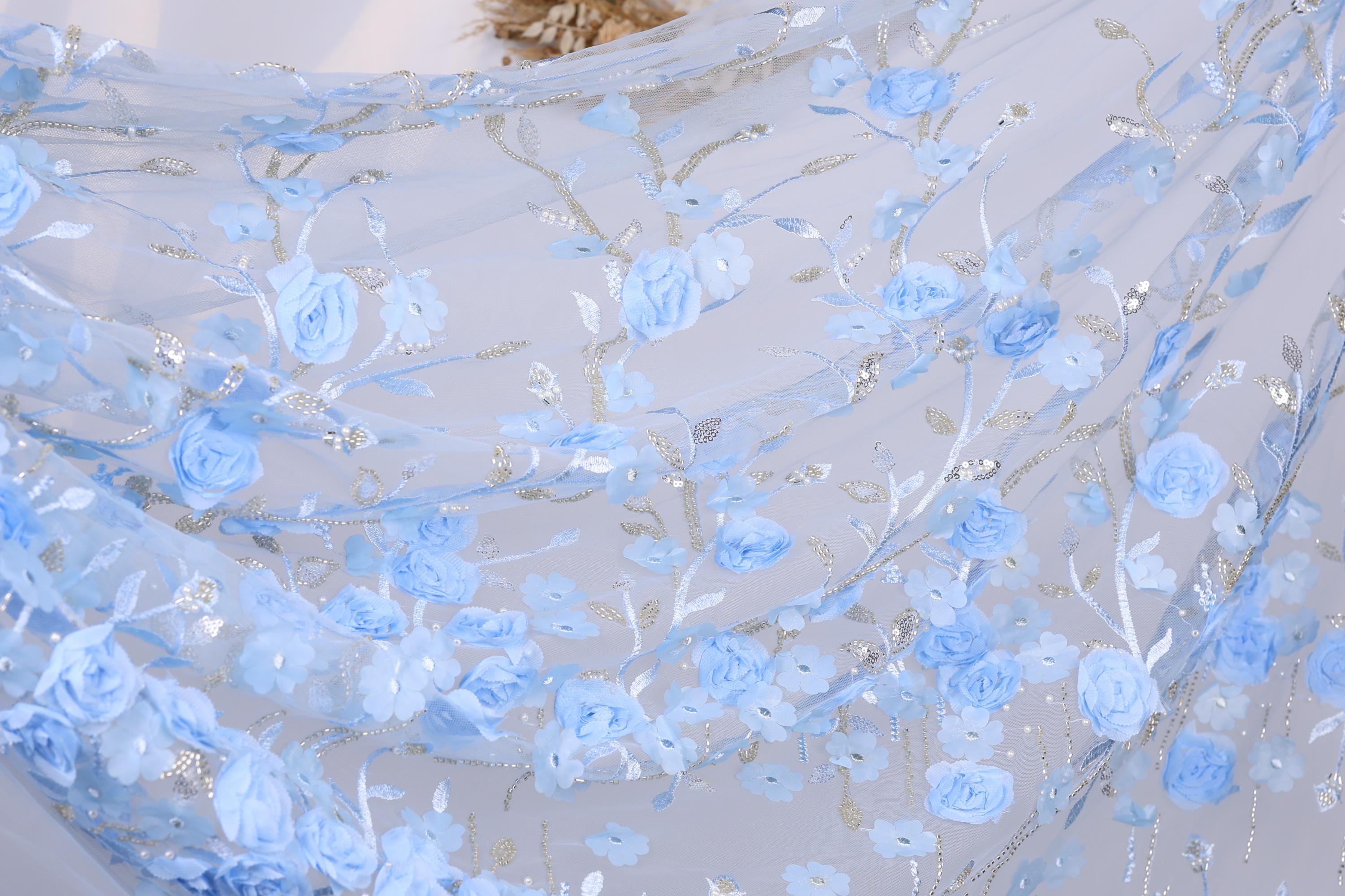 a close-up view of a delicate, light blue fabric adorned with a floral pattern of blue flowers and leaves. The fabric appears to be made of sheer material, giving it a soft and airy appearance. The intricate design and the delicate nature of the flowers and leaves create a visually appealing and elegant look.