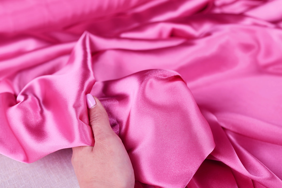 A hand is holding a piece of pink satin fabric, which appears to be soft and smooth.