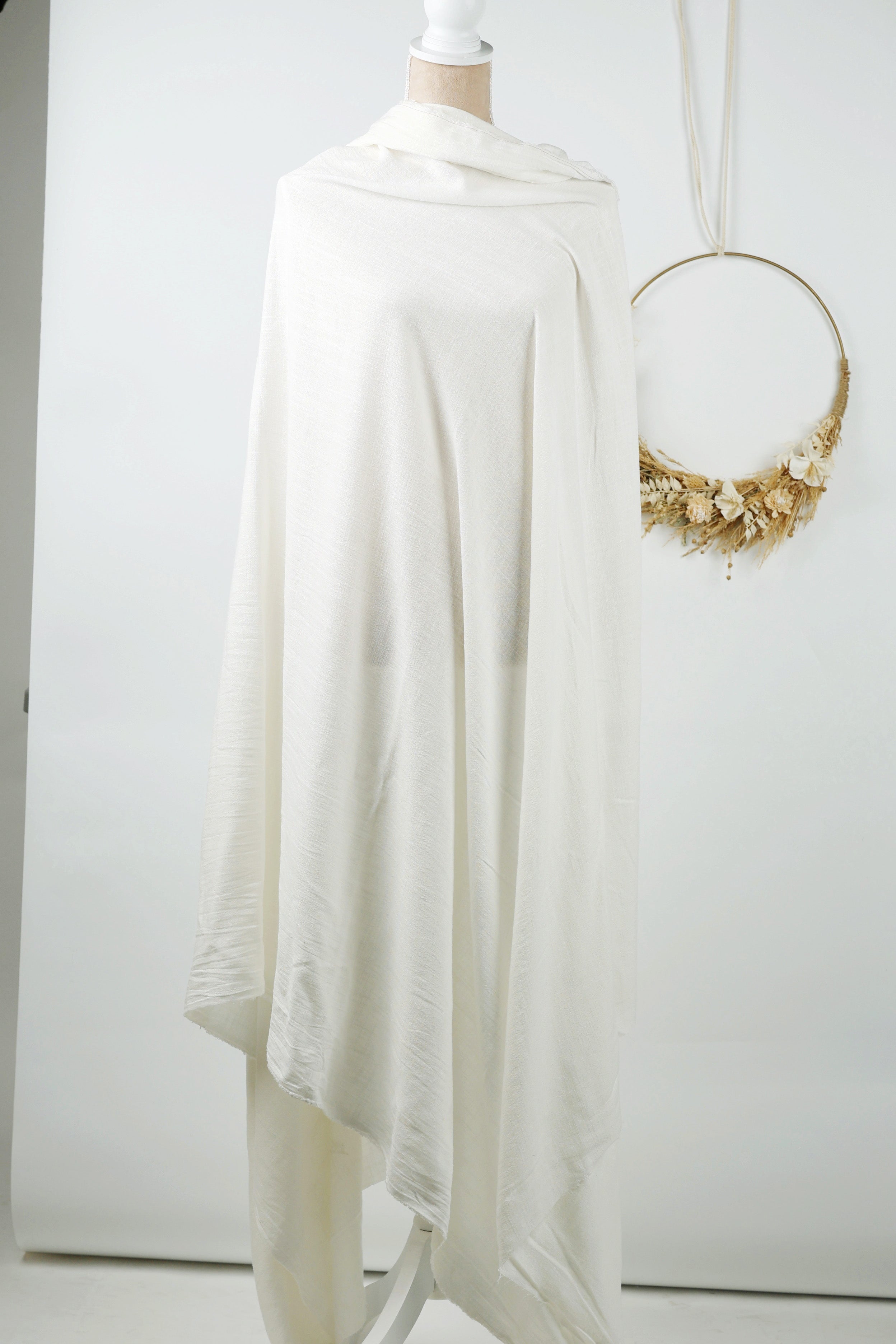 A white draped garment is displayed on a mannequin, with a gold wreath hanging on the wall behind it.