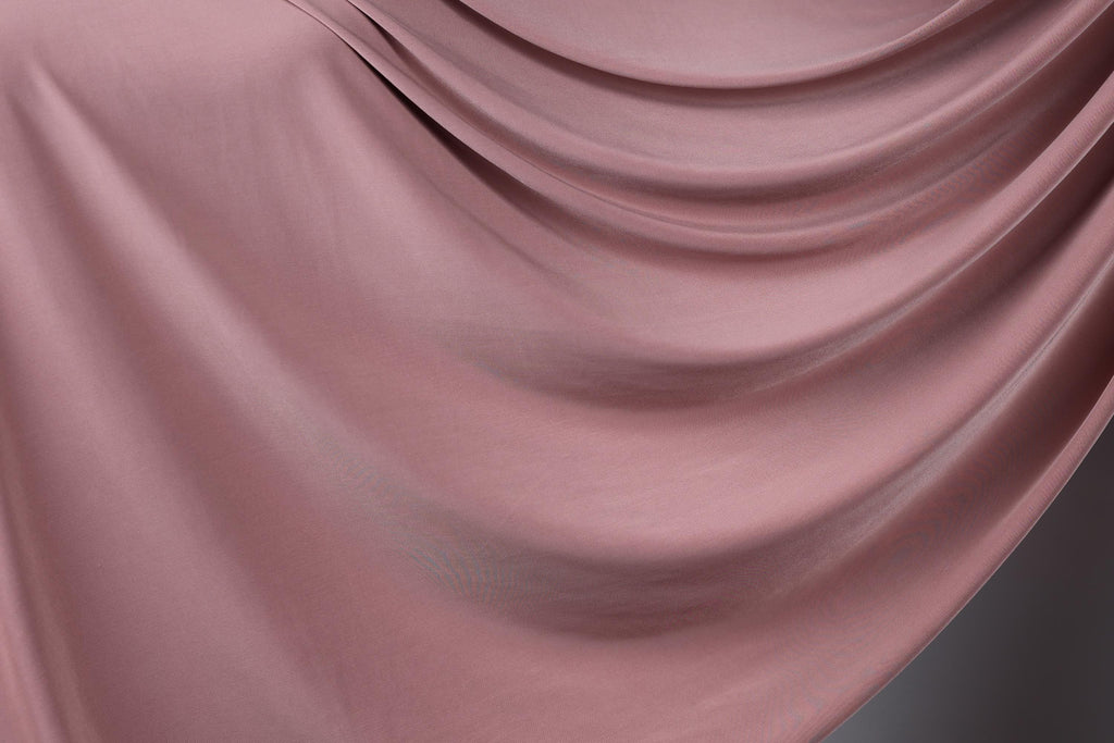 a close-up view of a soft, flowing pink fabric, possibly a satin or velvet material. The fabric has a smooth, shiny texture and is draped in a way that creates a sense of movement and elegance.