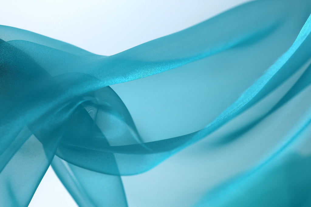 The image is a close-up of a flowing, turquoise-colored fabric, with the fabric appearing to be made of silk or a similar material. The fabric has a smooth, shiny texture and is captured in a dynamic, flowing pose, creating a sense of movement and elegance.