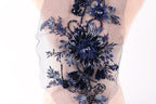 a close-up view of a delicate, intricate floral embroidery on a fabric. The embroidery is composed of various blue and black flowers and leaves, creating a visually striking and detailed design. The fabric appears to be a sheer material, which allows the embroidery to be clearly visible. The image does not contain any text or other objects, and the focus is solely on the embroidered floral design.