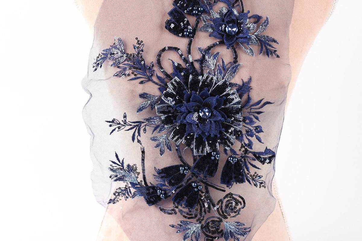 a close-up view of a delicate, intricate floral embroidery on a fabric. The embroidery is composed of various blue and black flowers and leaves, creating a visually striking and detailed design. The fabric appears to be a sheer material, which allows the embroidery to be clearly visible. The image does not contain any text or other objects, and the focus is solely on the embroidered floral design.