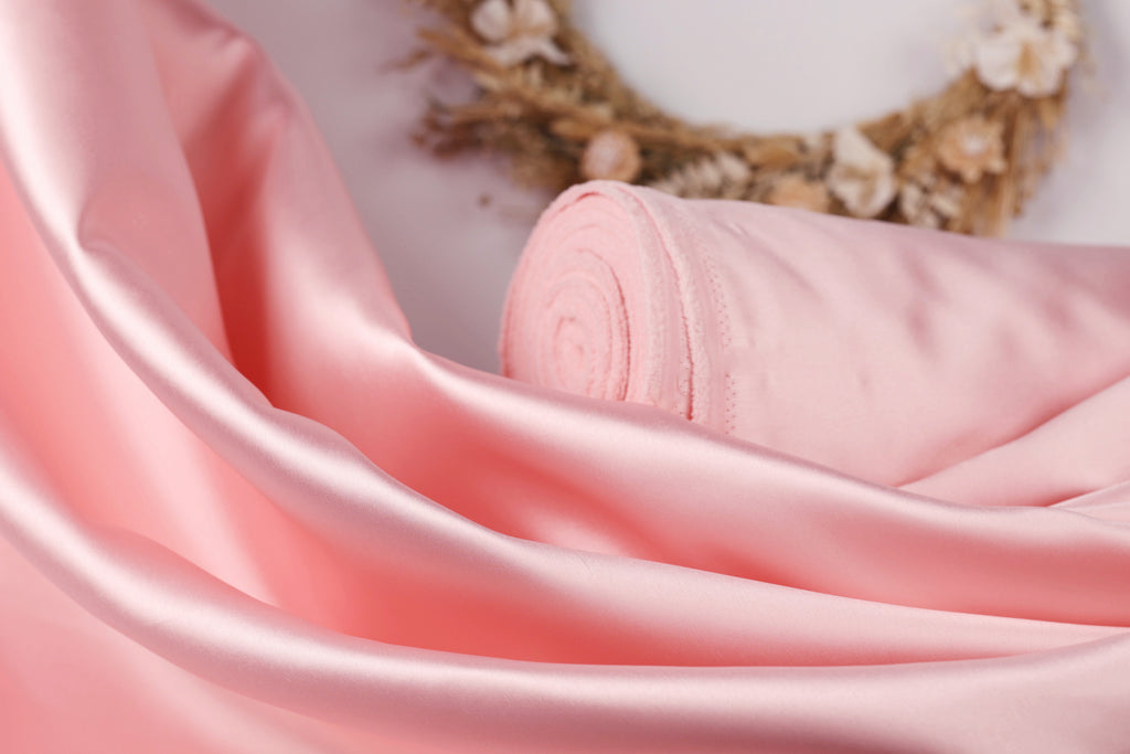 A close-up image of a soft, pink satin fabric, with a wreath of flowers visible in the background.