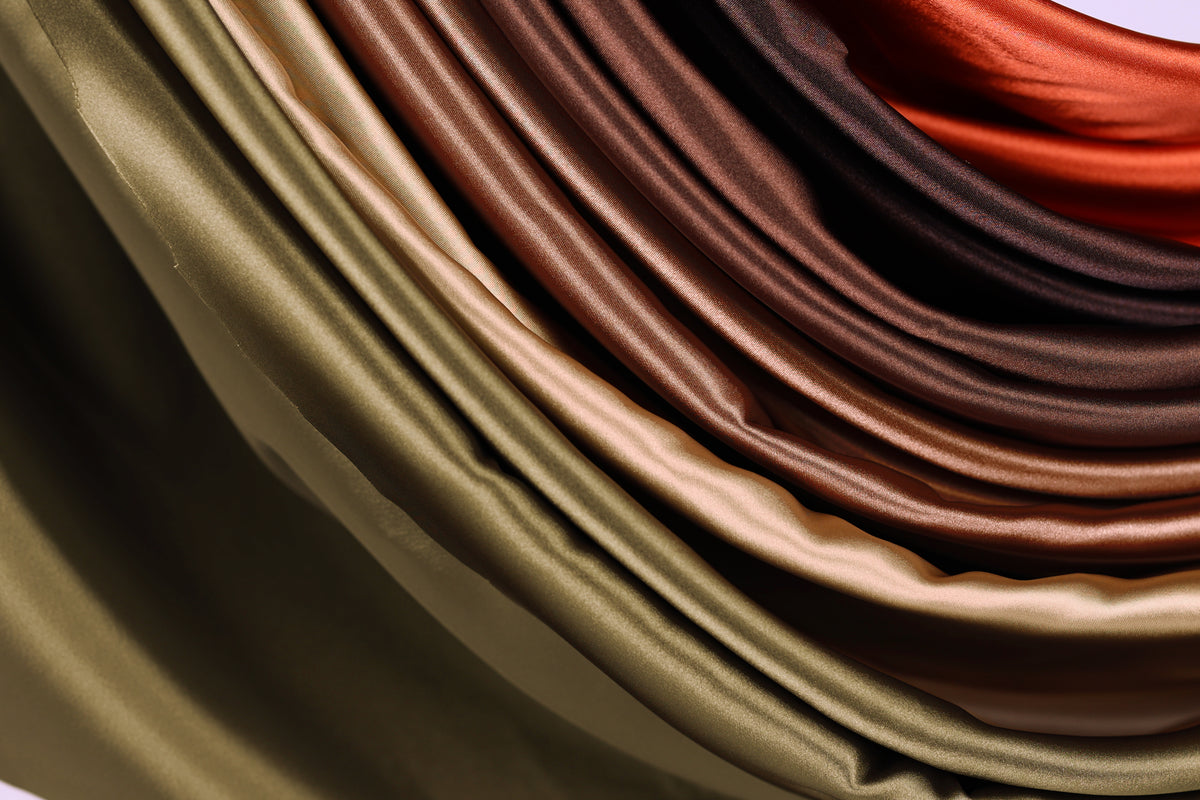 A close-up view of a pile of colorful satin fabric, with a gradient of colors ranging from green to brown and orange.