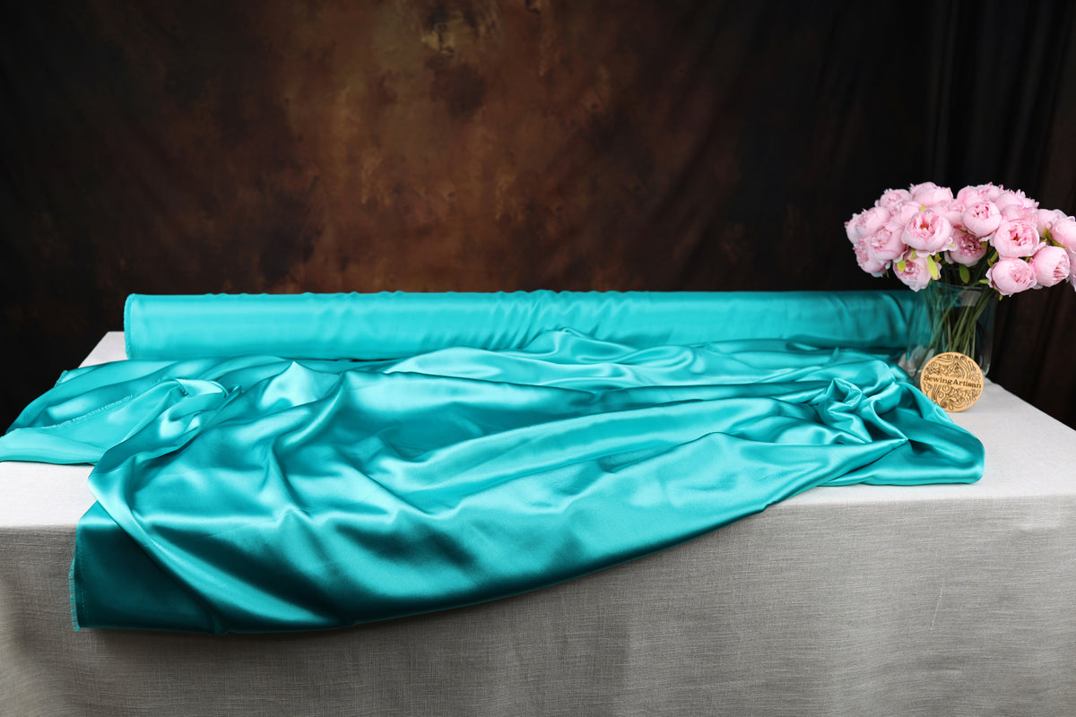 a turquoise satin fabric draped over a white tablecloth, with a vase of pink roses placed next to it.