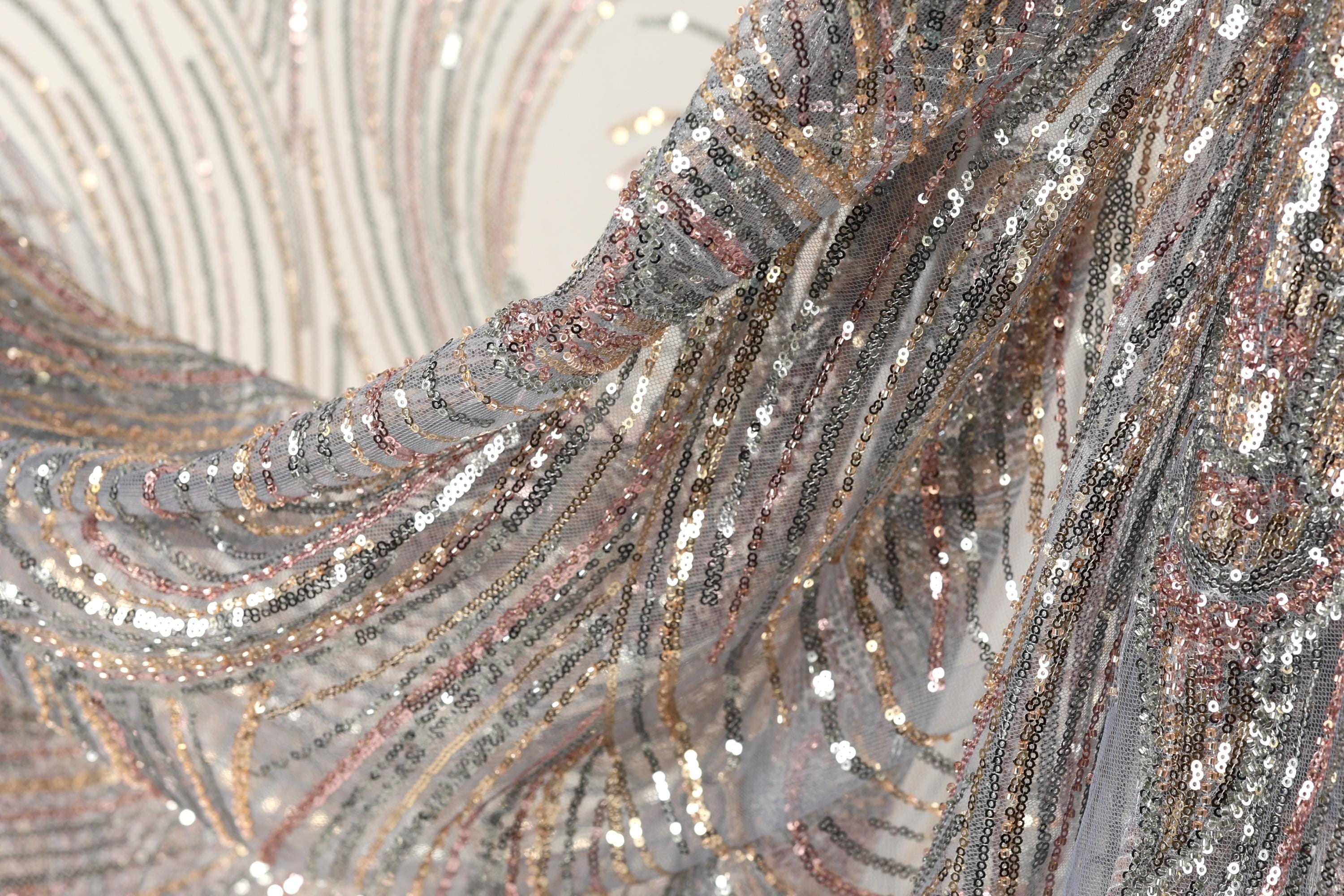 a close-up view of a fabric with a shimmering, sequined texture in shades of silver, gold, and pink. The fabric appears to be made of a sheer material, giving it a delicate and elegant appearance.