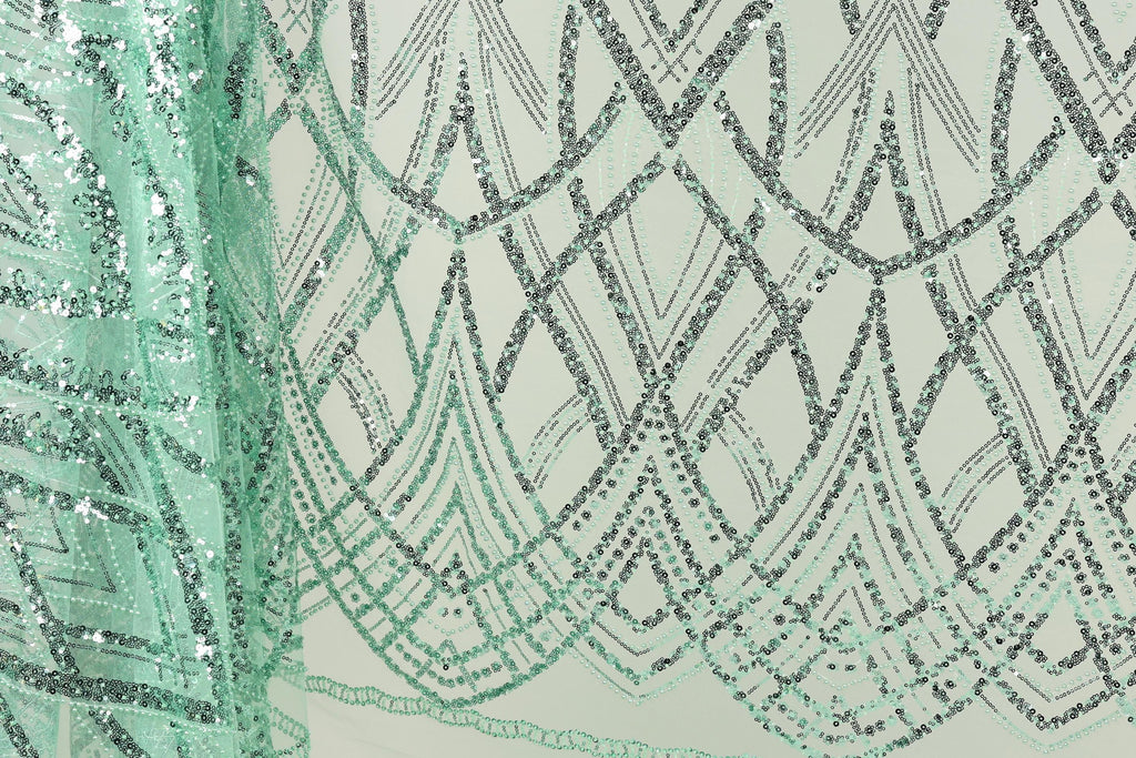 a close-up view of a fabric with a repeating pattern of green and black sequin designs. The pattern appears to be a geometric design with intersecting lines and curves, creating a visually striking and intricate design.