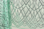 a close-up view of a fabric with a repeating pattern of green and black sequin designs. The pattern appears to be a geometric design with intersecting lines and curves, creating a visually striking and intricate design.
