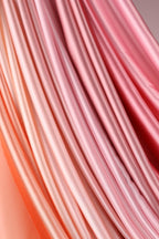 a close-up view of a soft, flowing fabric with a gradient of pink and orange hues. The fabric appears to be made of silk or satin, and it is arranged in a way that creates a sense of movement and texture. The image does not contain any discernible objects or actions, and it does not depict a scene or a person. The focus is solely on the fabric and its colors.