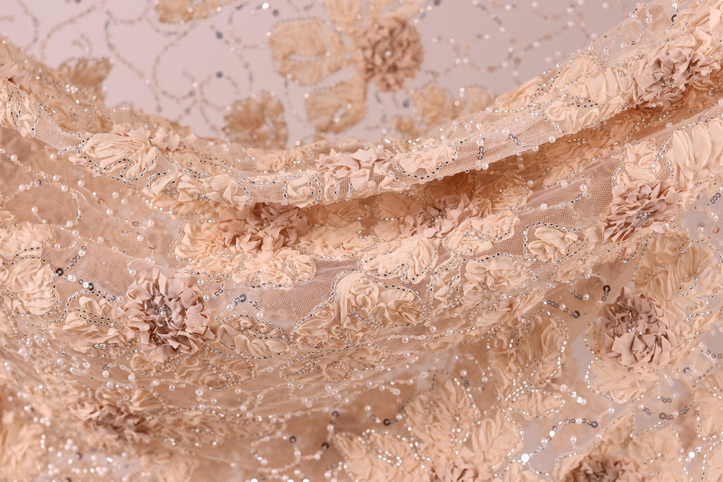 The image is a close-up of a delicate, intricately embroidered fabric with a floral pattern. The fabric appears to be made of a light-colored material, possibly lace or chiffon, with a floral design that includes various shades of pink and beige flowers and leaves. The fabric is intricately detailed, with the individual flowers and leaves appearing to be made of small beads or sequins, adding a touch of sparkle and texture to the overall design.