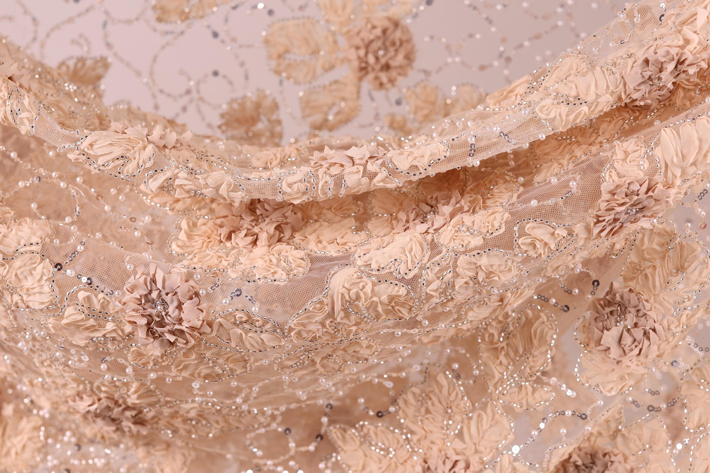 The image is a close-up of a delicate, intricately embroidered fabric with a floral pattern. The fabric appears to be made of a light-colored material, possibly lace or chiffon, with a floral design that includes various shades of pink and beige flowers and leaves. The fabric is intricately detailed, with the individual flowers and leaves appearing to be made of small beads or sequins, adding a touch of sparkle and texture to the overall design.