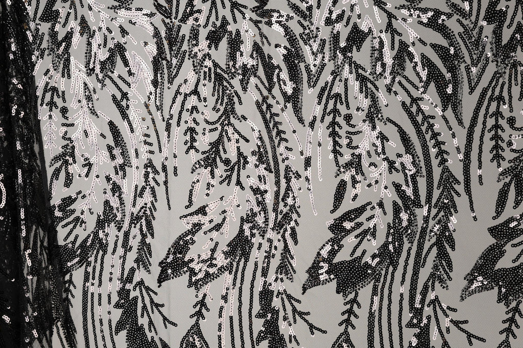a close-up view of a black and white patterned fabric with a repeating design of black and white sequins or beads. The pattern appears to be a floral or leaf-like design, with the sequins or beads arranged in a way that creates a sense of movement and texture. The fabric has a textured surface, and the black and white color scheme adds a classic and timeless aesthetic to the design.