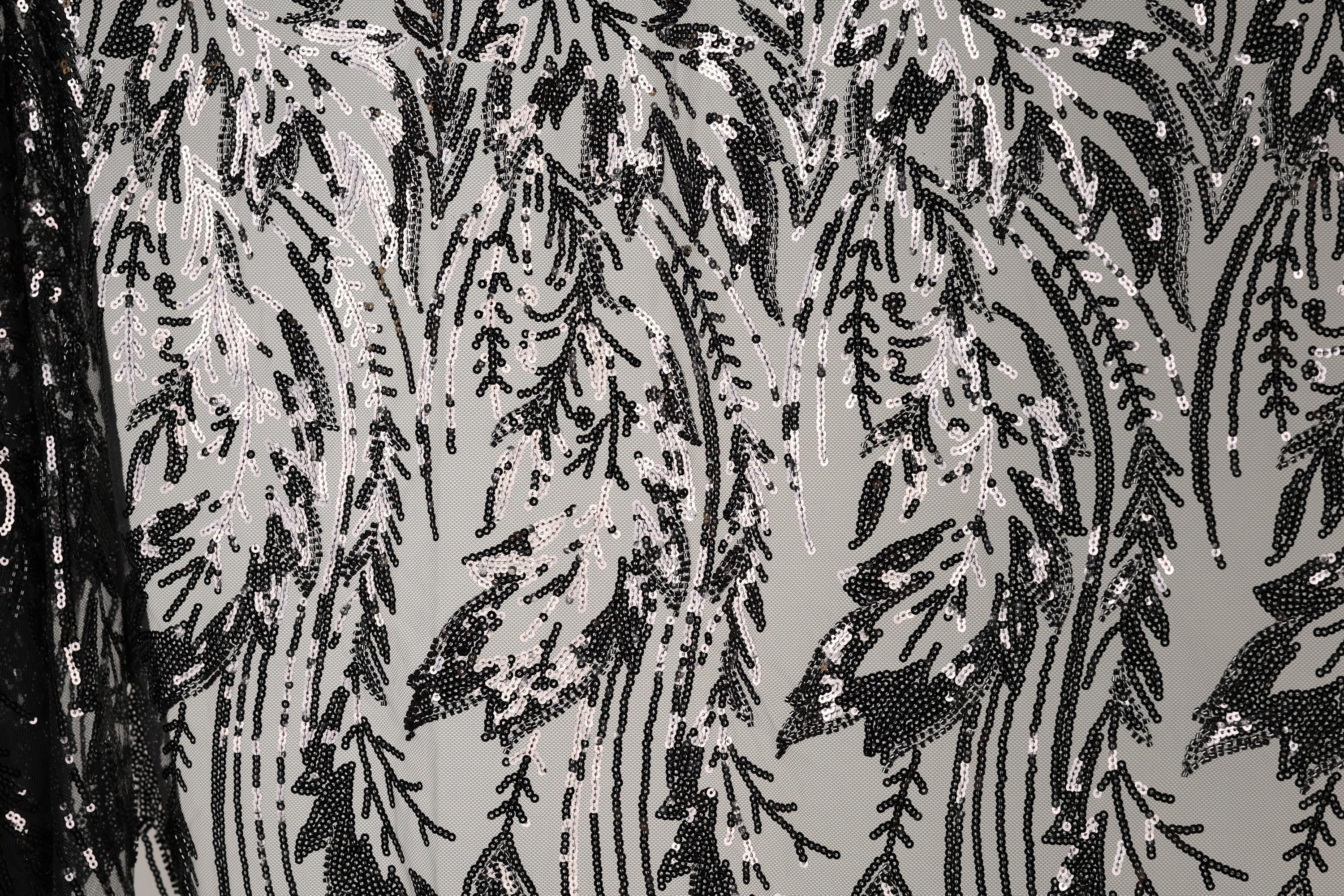 a close-up view of a black and white patterned fabric with a repeating design of black and white sequins or beads. The pattern appears to be a floral or leaf-like design, with the sequins or beads arranged in a way that creates a sense of movement and texture. The fabric has a textured surface, and the black and white color scheme adds a classic and timeless aesthetic to the design.