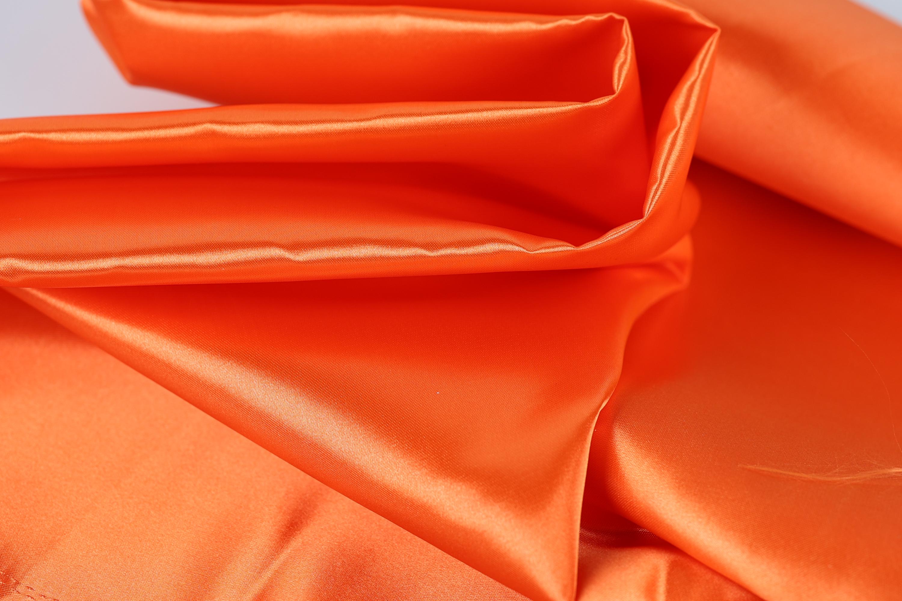 The image is a close-up of a bright orange fabric, possibly silk or satin, with a shiny and smooth texture.