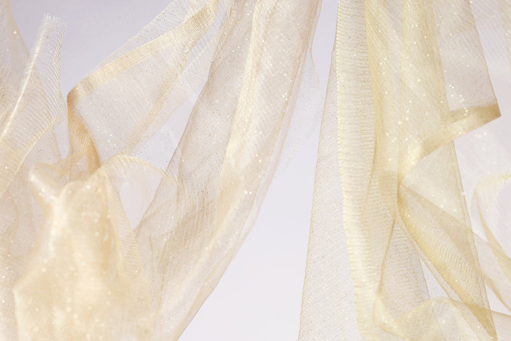 a close-up view of a delicate, flowing fabric with a shimmering, golden-yellow hue. The fabric appears to be made of sheer material, giving it a soft and delicate appearance. The background is a plain white color, which contrasts with the fabric and draws attention to it. The image does not contain any discernible text or countable objects, and the relative positions of the objects are not clear, making it difficult to determine the specific details of the image.