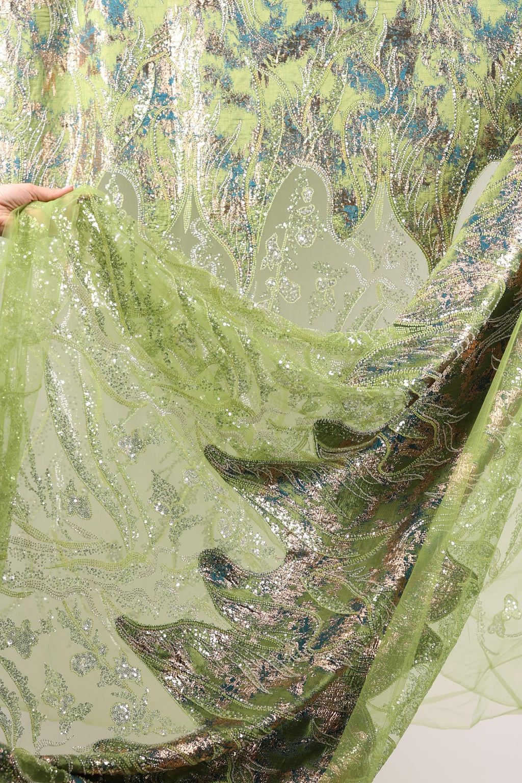 a close-up view of a fabric with a green and gold pattern, with a hand holding it up to reveal the intricate details.