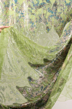 a close-up view of a fabric with a green and gold pattern, with a hand holding it up to reveal the intricate details.
