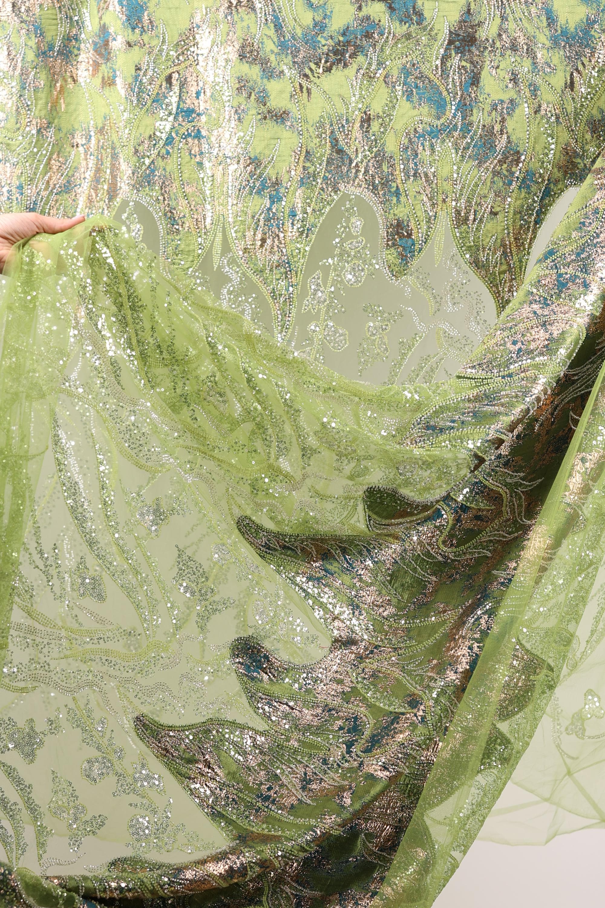 a close-up view of a fabric with a green and gold pattern, with a hand holding it up to reveal the intricate details.