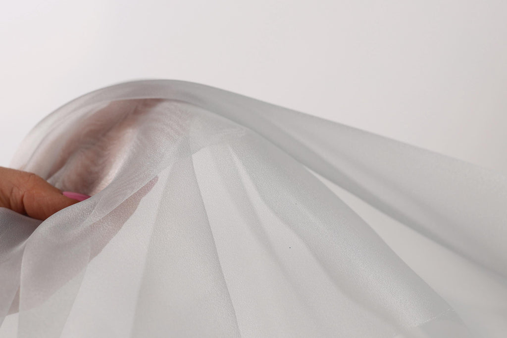 A hand is holding a white fabric, which appears to be a sheer or sheer-like material. The fabric is being held up against a plain white background, creating a simple and minimalist visual composition.