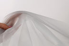 A hand is holding a white fabric, which appears to be a sheer or sheer-like material. The fabric is being held up against a plain white background, creating a simple and minimalist visual composition.