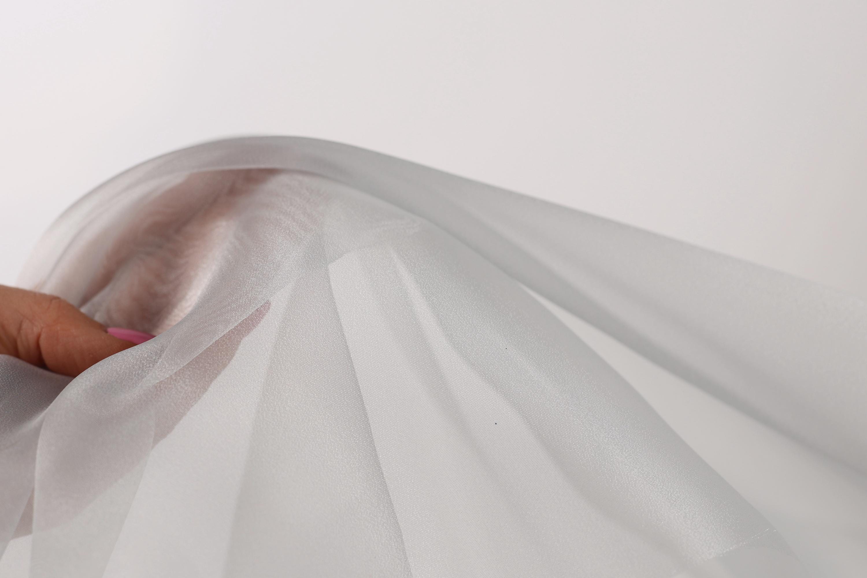 A hand is holding a white fabric, which appears to be a sheer or sheer-like material. The fabric is being held up against a plain white background, creating a simple and minimalist visual composition.