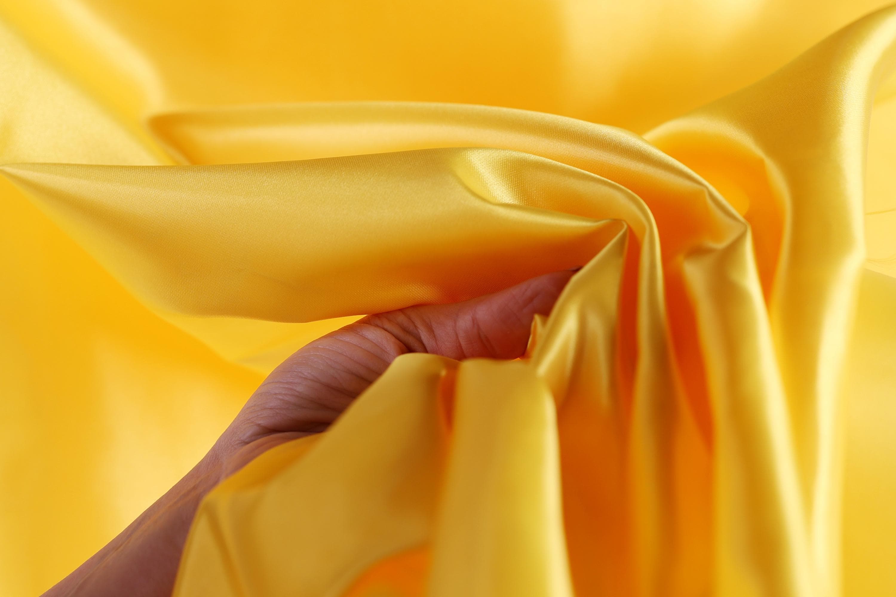 A hand is holding a yellow fabric, which appears to be a soft, flowing material. The fabric is being held up against a bright yellow background, creating a visually striking contrast.