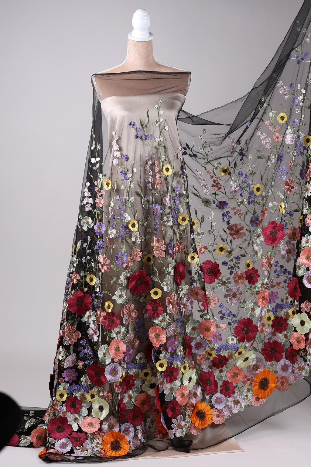 A mannequin wearing a long, flowing dress with a sheer overlay featuring a vibrant floral pattern in various colors, including red, orange, yellow, and purple.